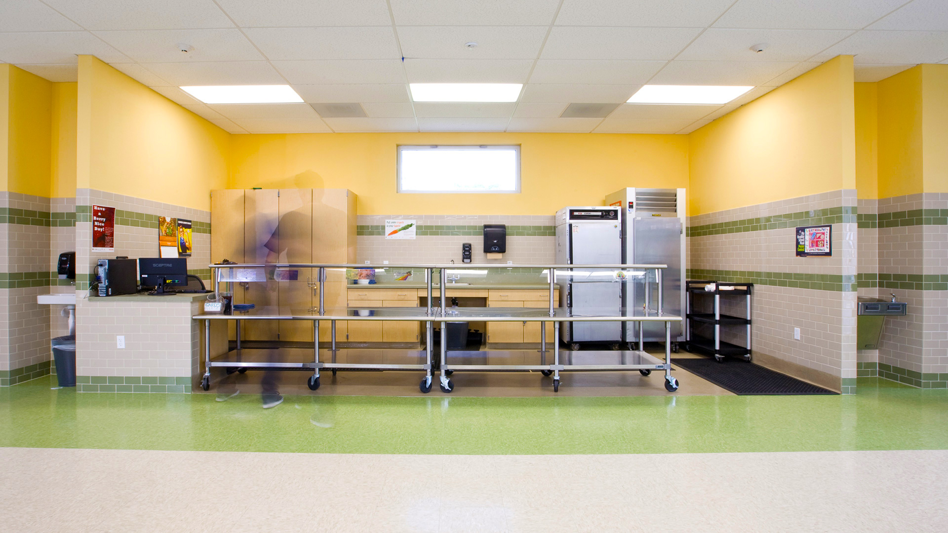 Interior of multi-use room looking at kitchen area and serving counter, with green vinyl flooring and yellow walls.