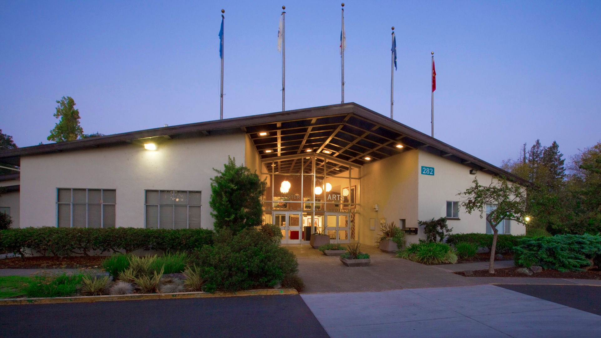 Front of Center for the Arts building at dusk, with glowing entrance and tall green bushes in front.