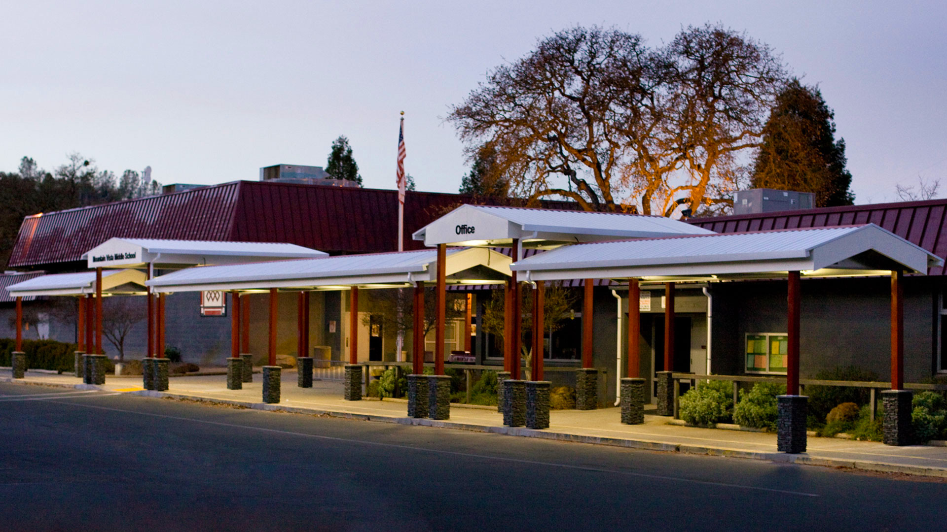 Metal shade structure at dusk, with red posts and white tops, with 'Office' lettered at the secondary entrance.