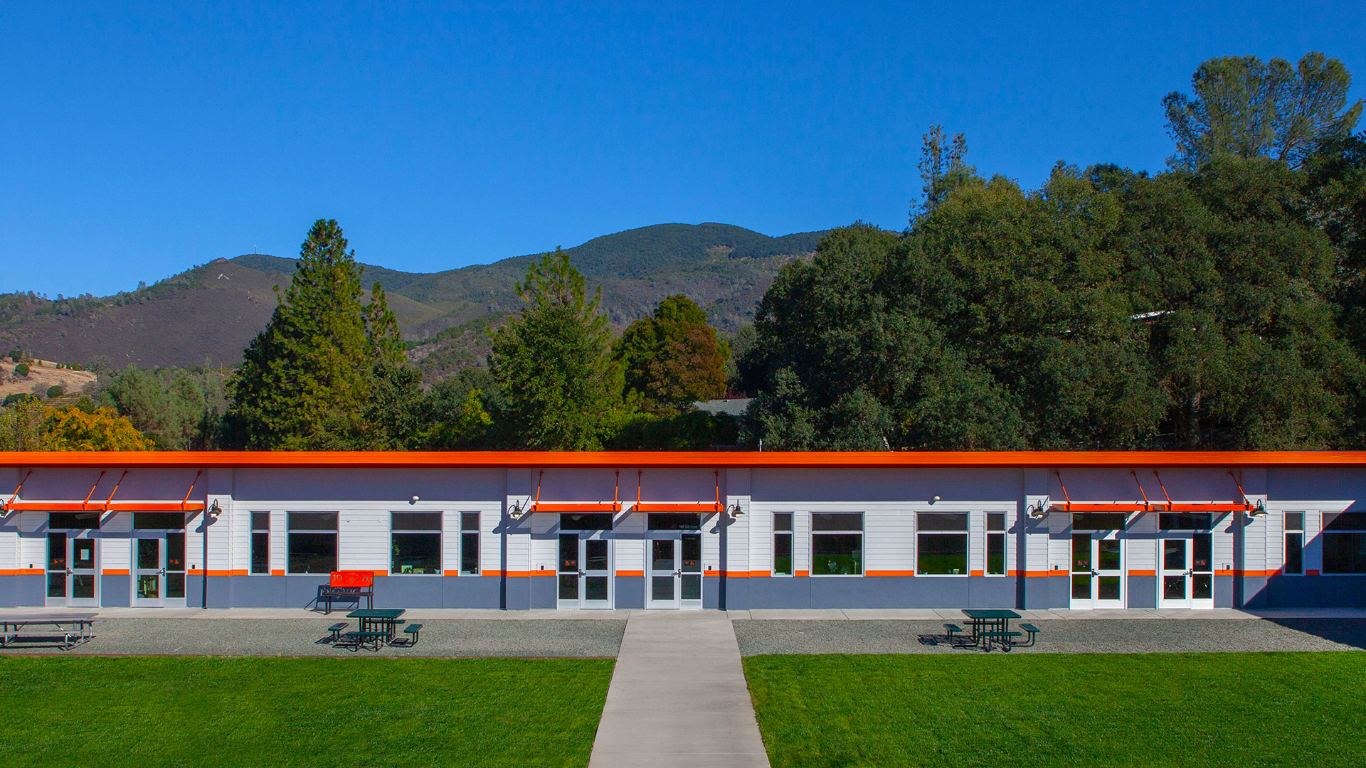 Wide shot of modular classroom wing. White walls with gray wainscot, orange trim and details. Mountains and grass around