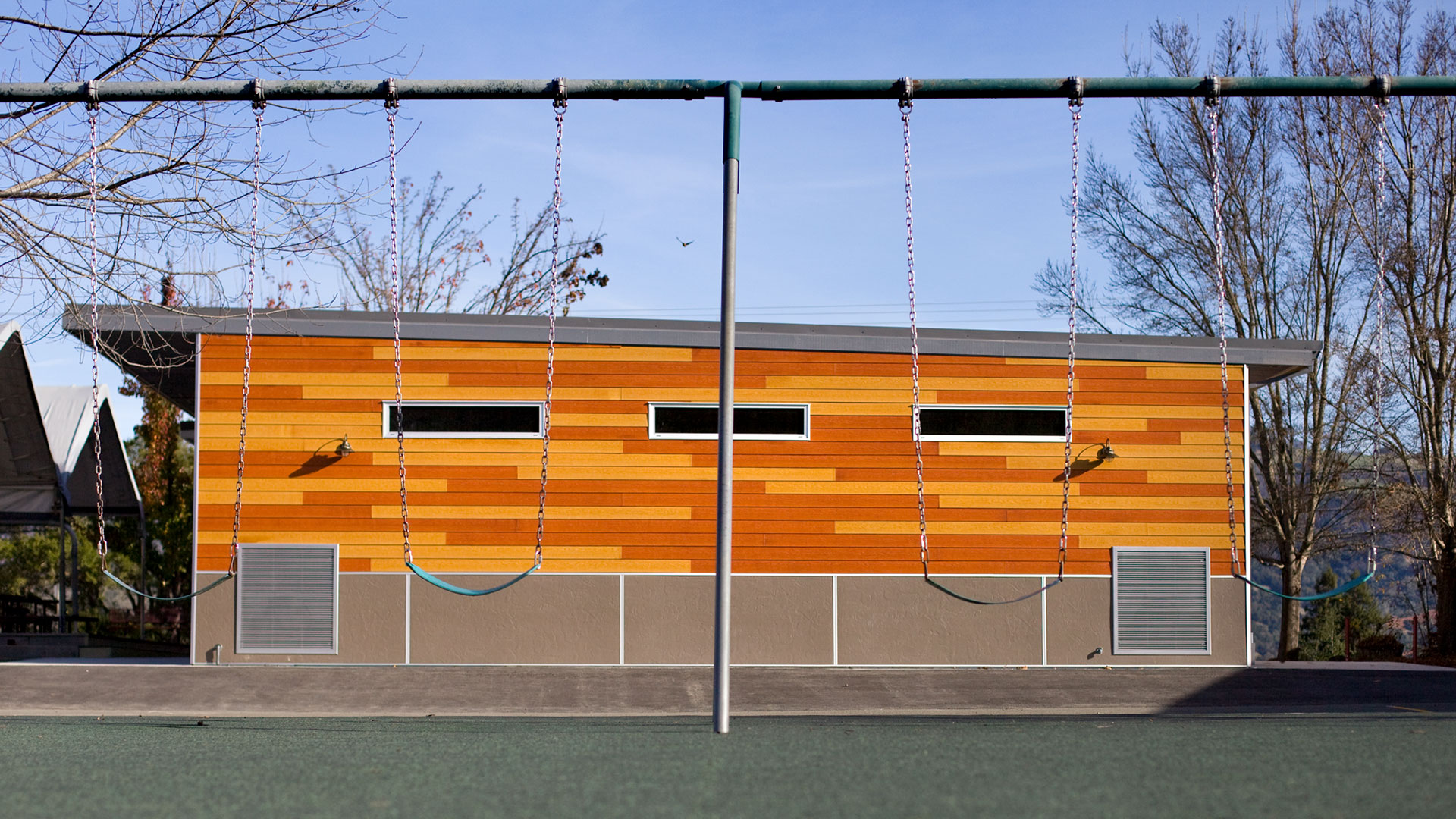 New multi-use room exterior, with wooden panel walls over gray wainscot, with playground swings in foreground.
