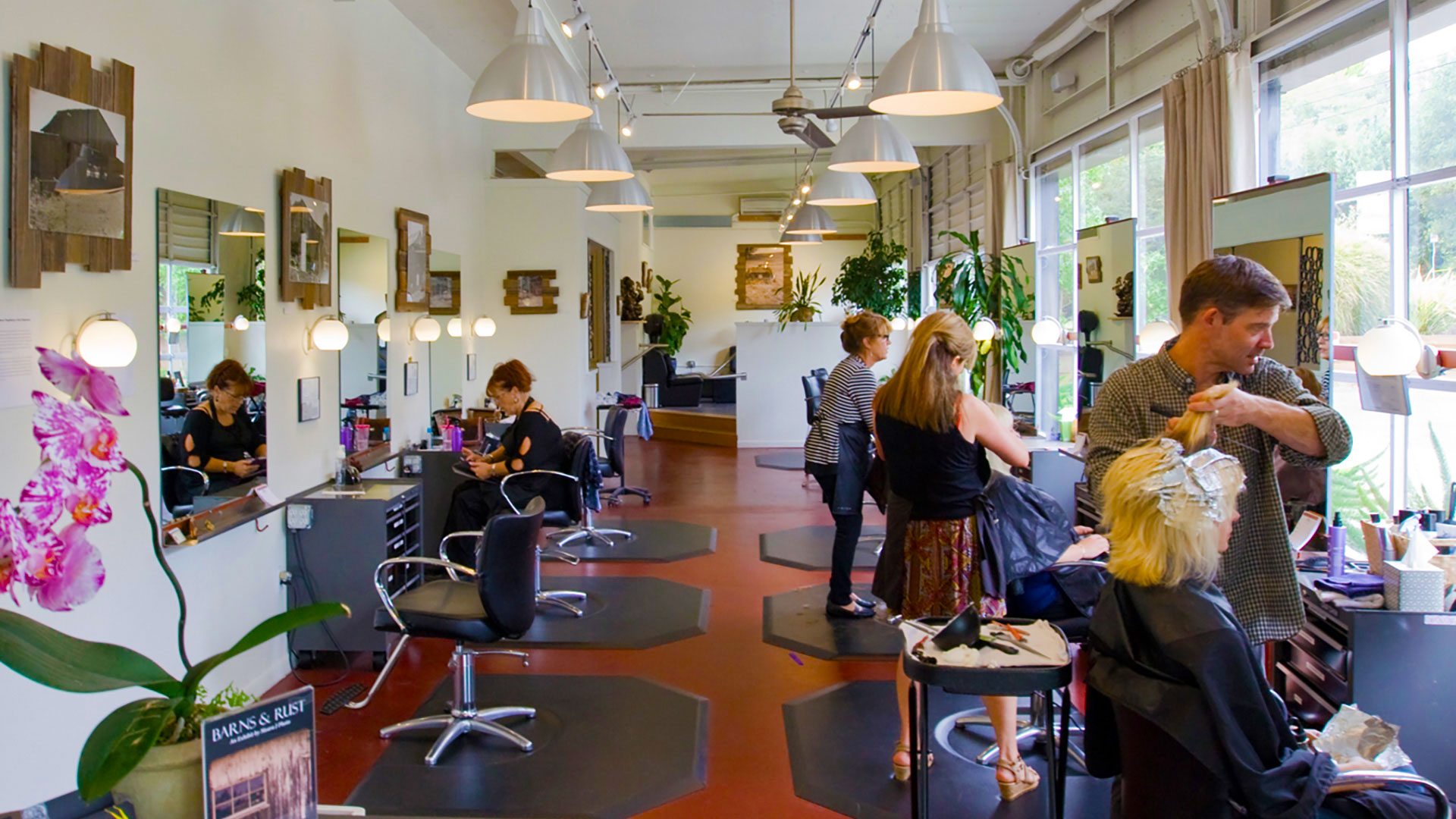 Interior of salon, salon chairs and mirrors stationed along red concrete floor and stylists working on customers.