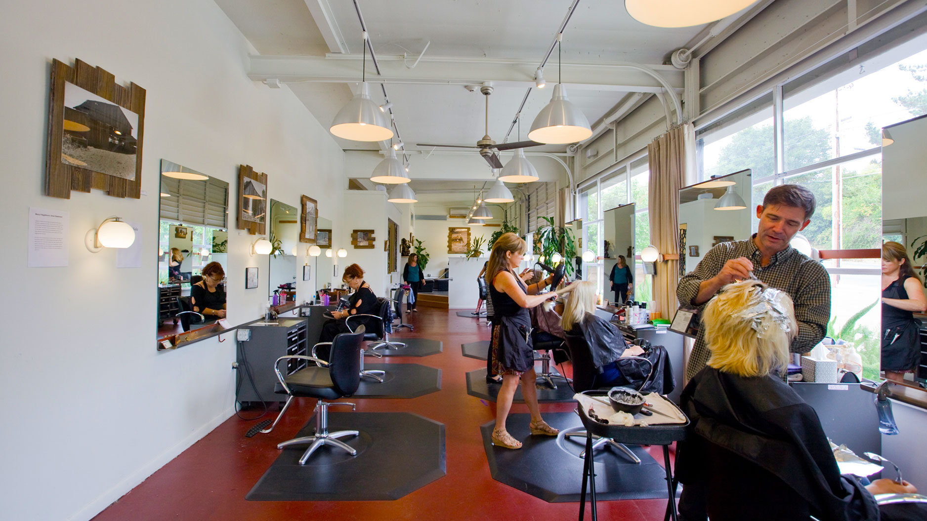 Interior of salon, salon chairs and mirrors stationed along red concrete floor and stylists working on customers.