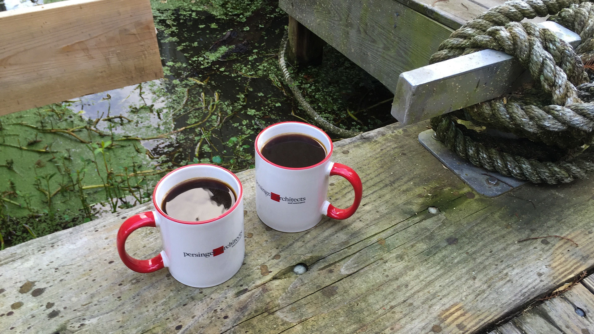 Pair of persinger mugs, filled with coffee, on a wooden dock with water below
