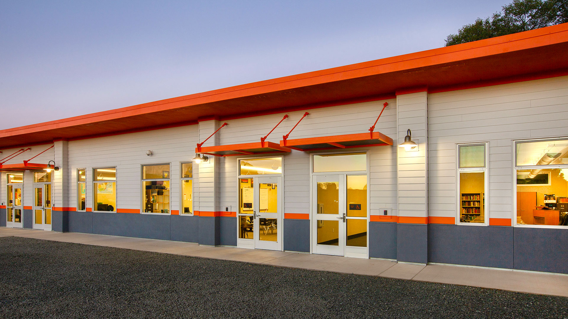 Row of modular classrooms, with white walls, gray wainscot, and orange details, roof, and awnings.