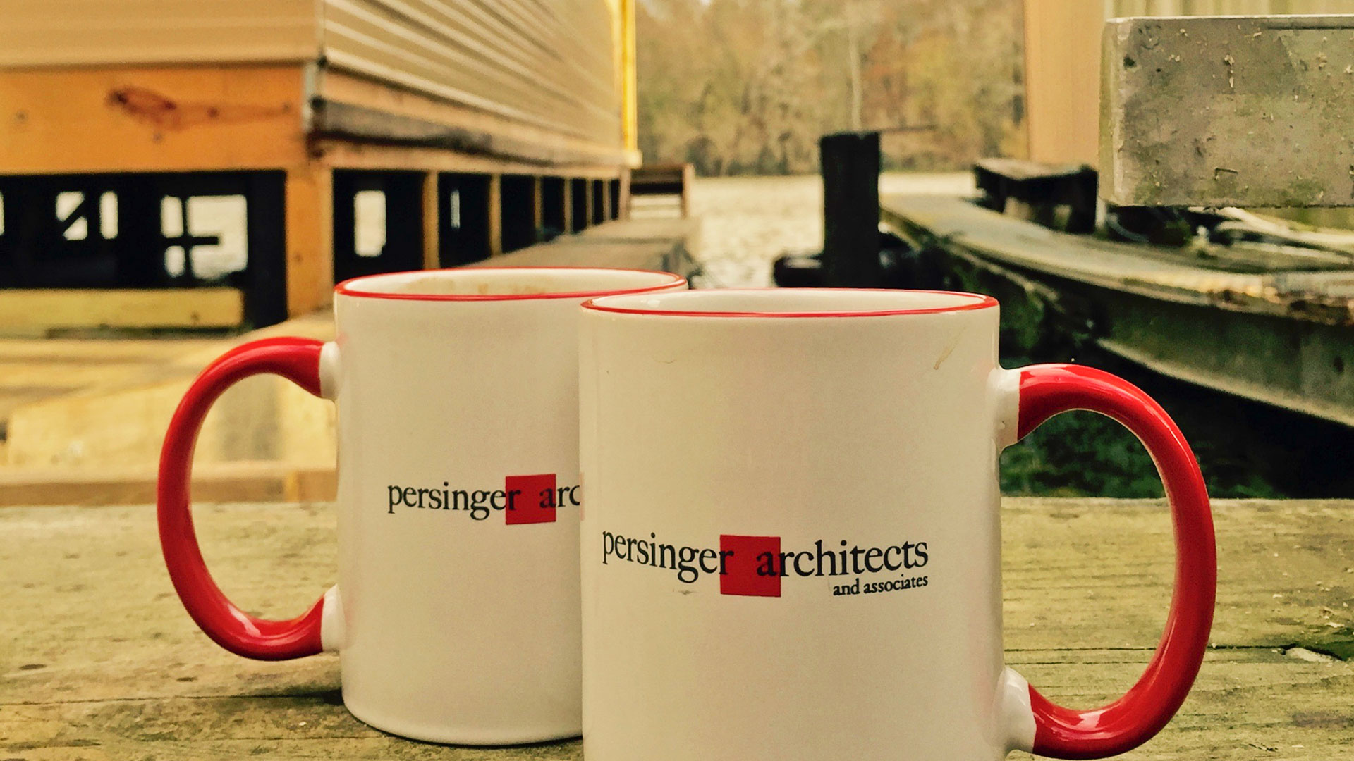 Pair of white mugs with persinger logo, red handles and lip, on a wooden dock with river and forest behind.