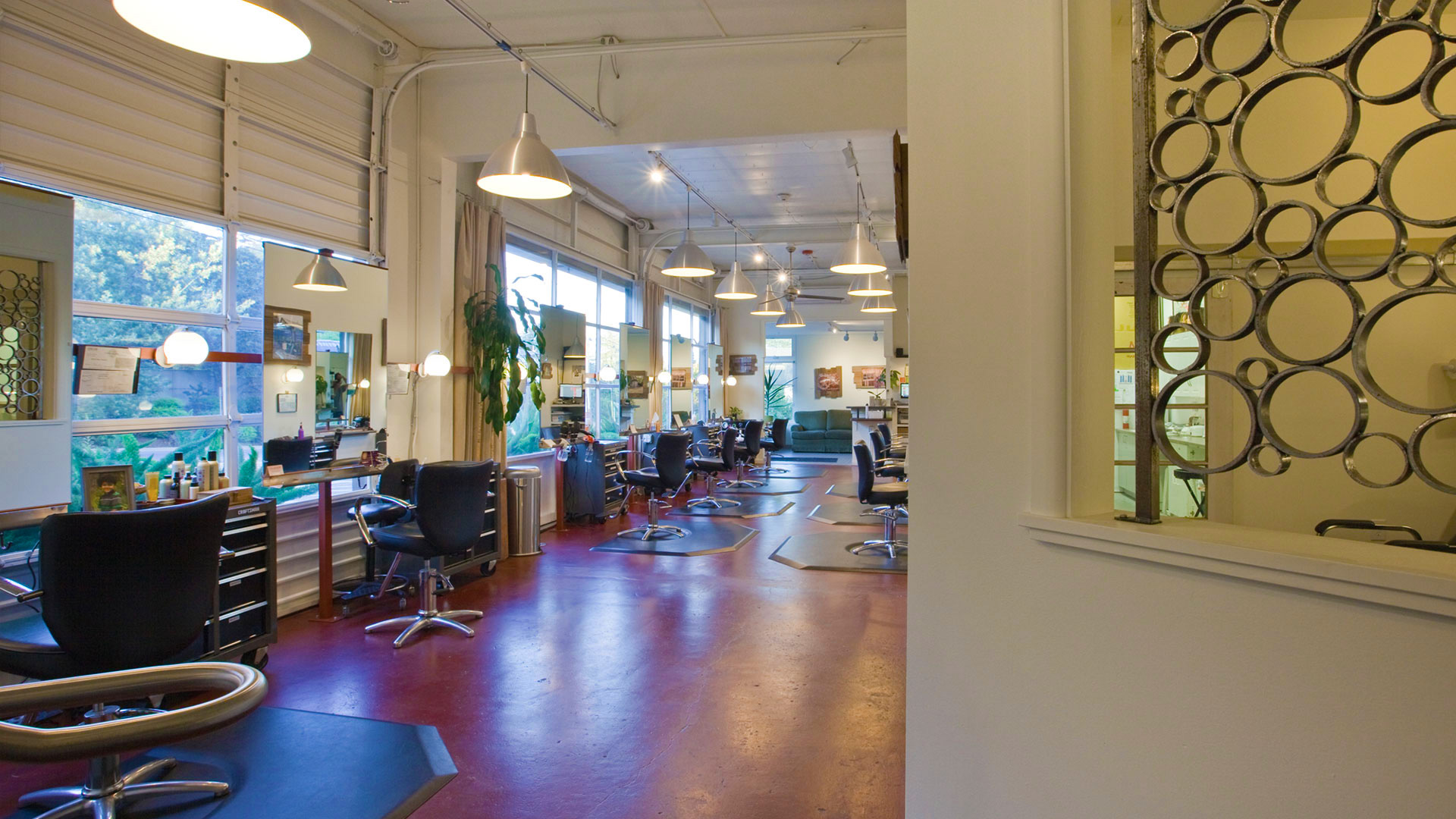 Interior of salon, salon chairs and mirrors stationed along red concrete floor, hanging pendent lights above.