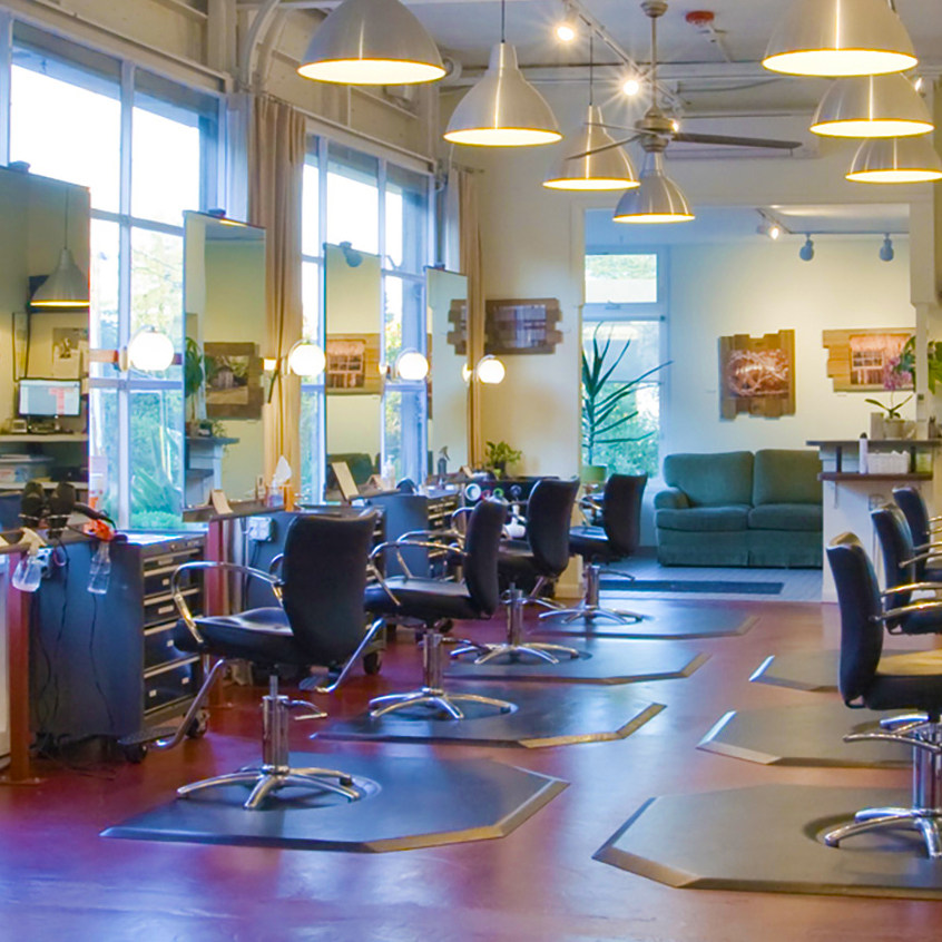 Interior of salon, salon chairs and mirrors stationed along red concrete floor, hanging pendent lights above.
