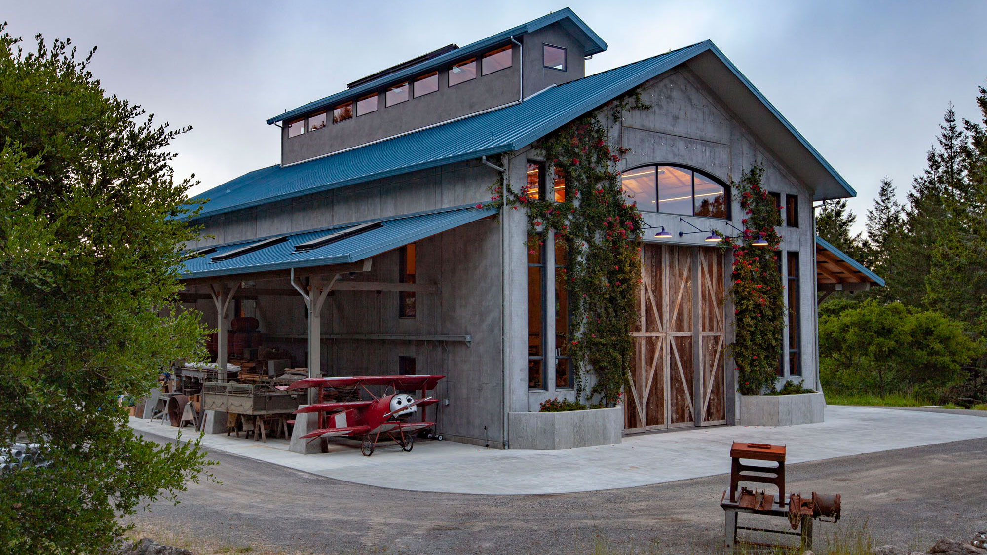 Side angle of concrete barn, double height and blue roof, with covered workspace adjacent.