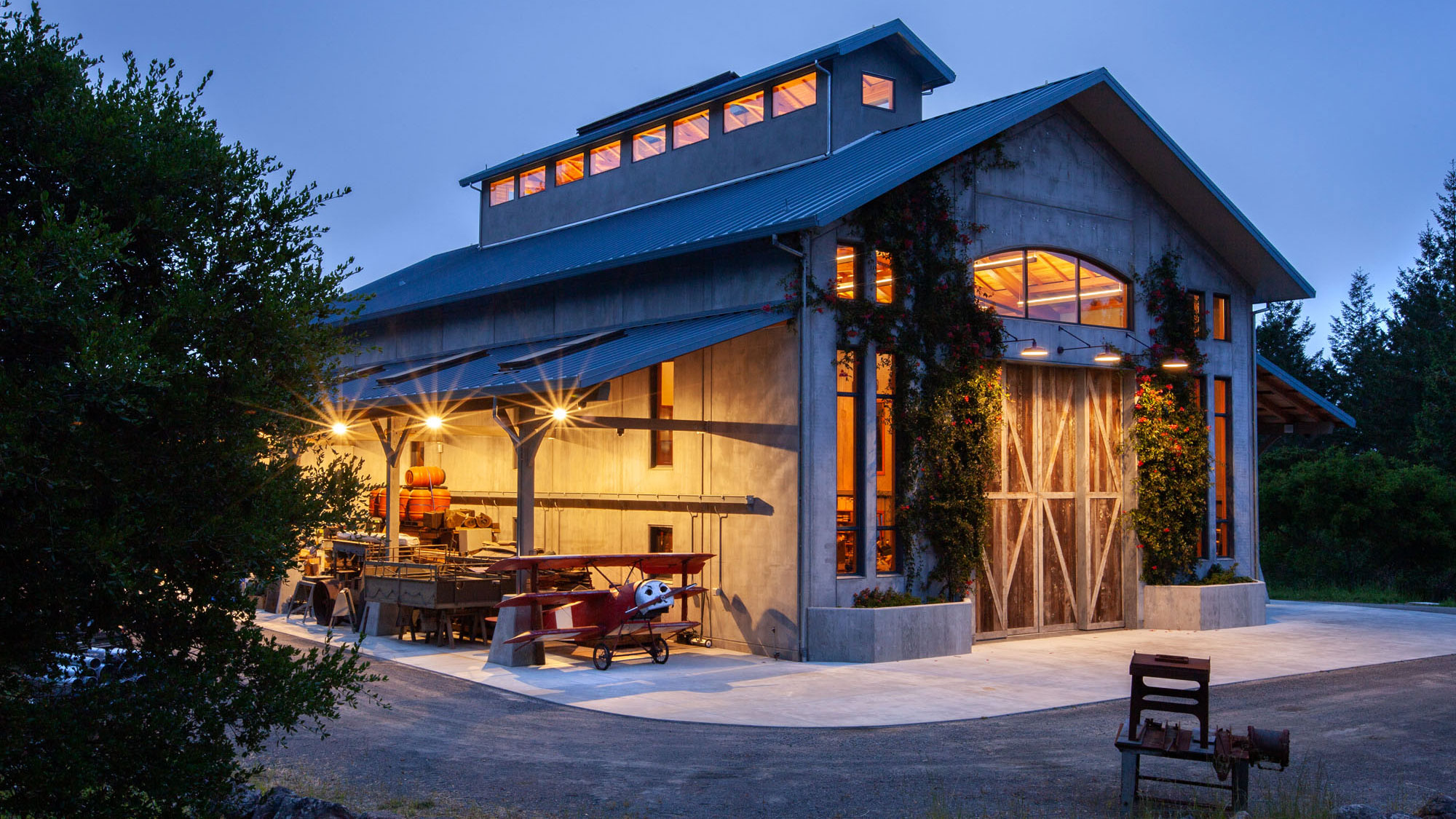 Side angle of concrete barn at night, double height and blue roof, with covered workspace adjacent and glowing windows.
