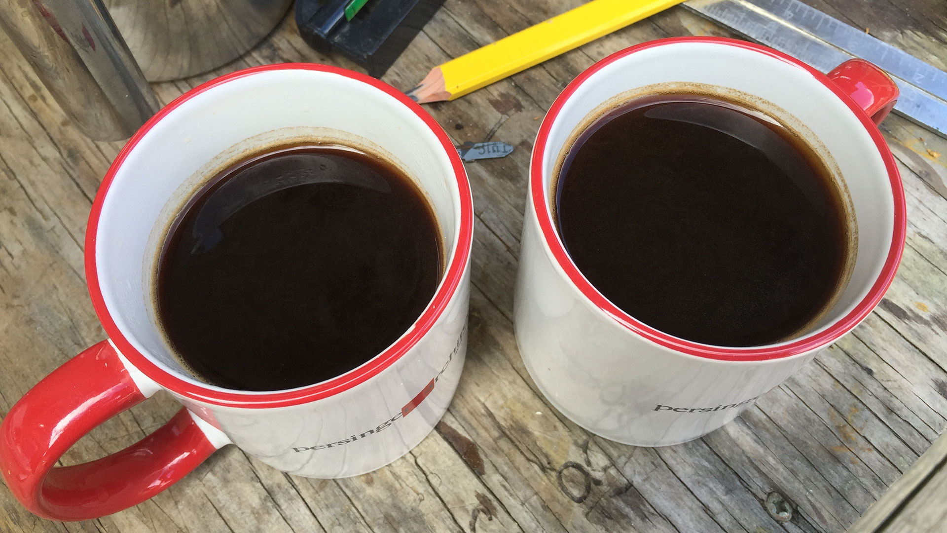 Pair of persinger mugs filled with coffee resting on wooden table with pencils around