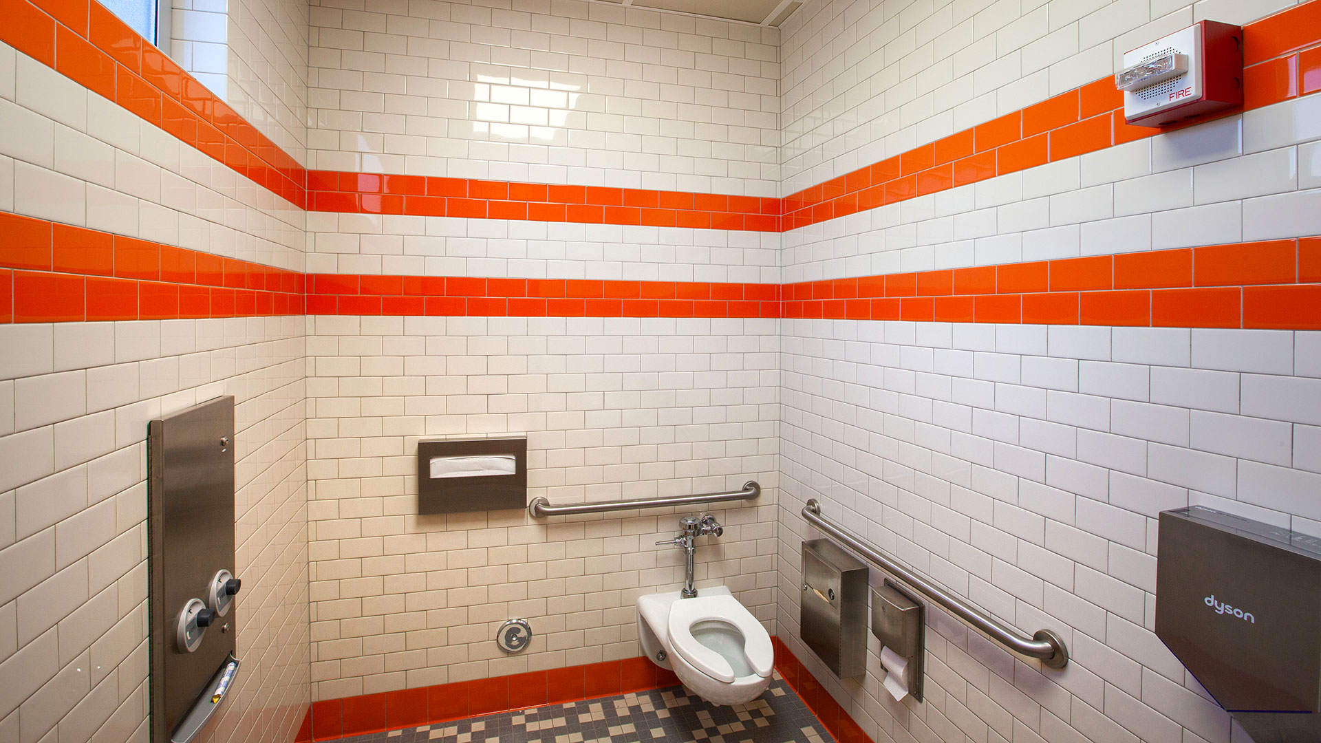 Interior of new restrooms, with white and orange tile walls and gray tile floors.