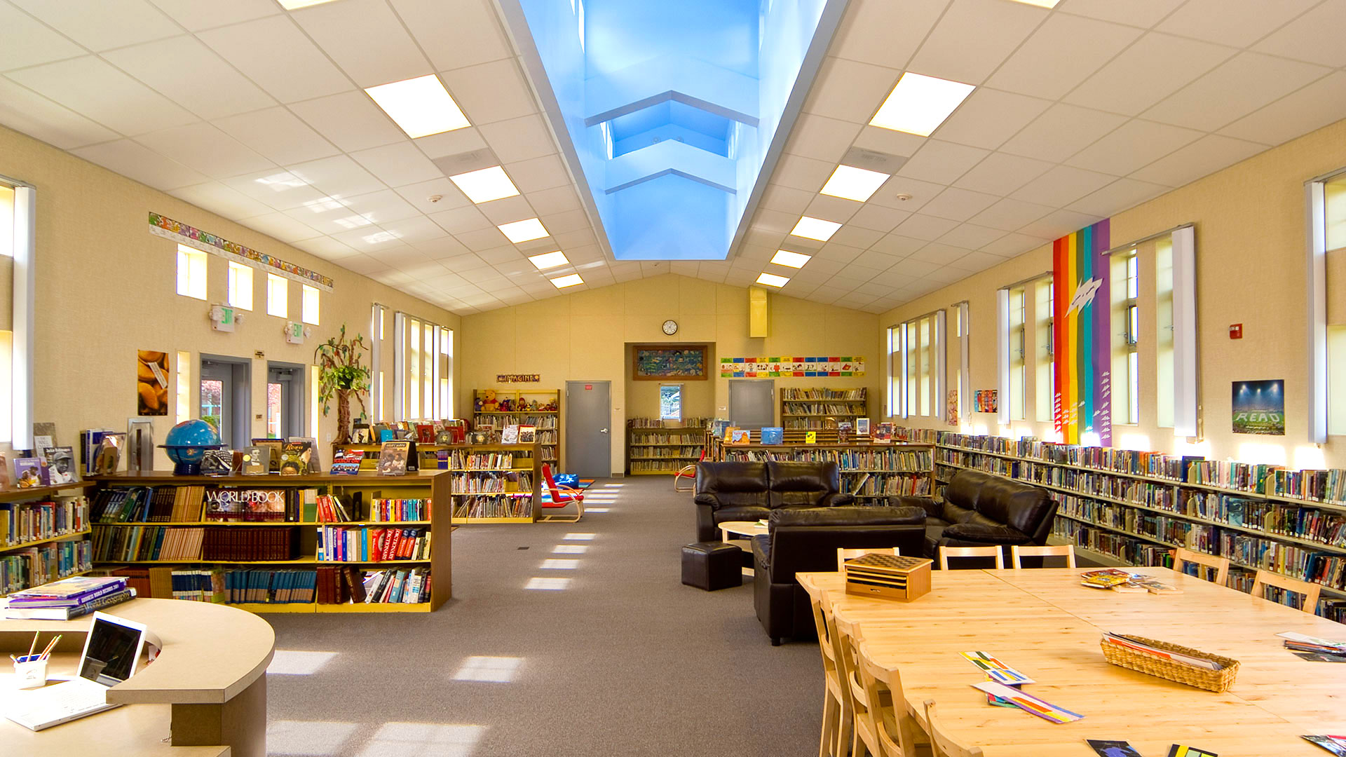 Library interior, looking across the building. Cushioned seating across from bookshelves and blue monitor above.