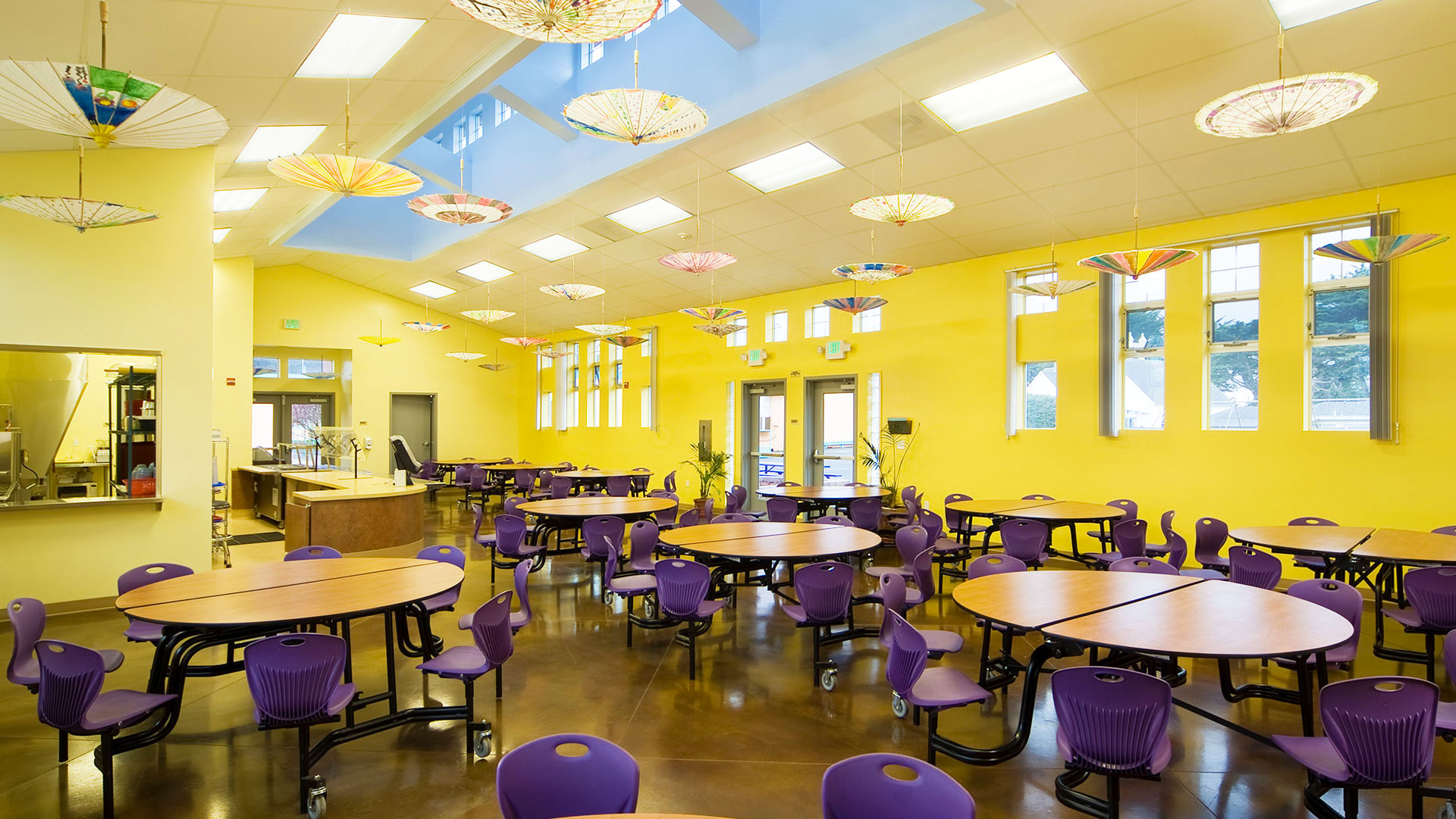 Interior of cafeteria, with bright yellow walls, circular tables and purple chairs.