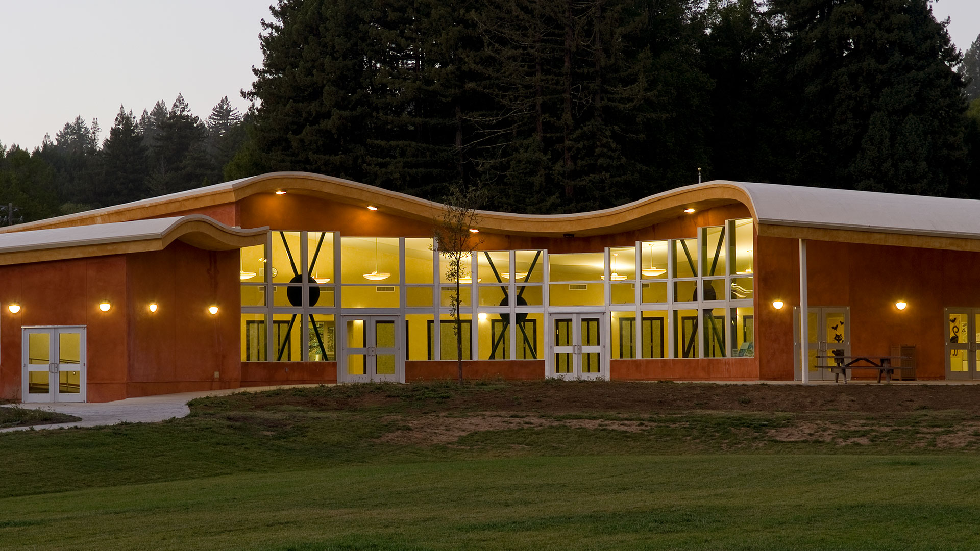 Exterior of school at dusk, with windows illuminated under the orange curving roof.