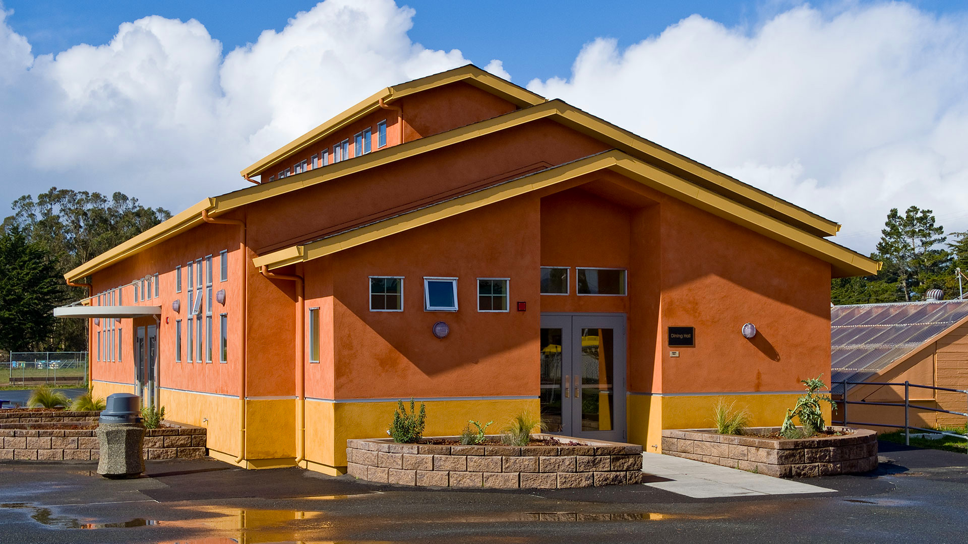 Exterior of completed cafeteria. Orange walls with a yellow wainscot and trim, with a black shingled roof.
