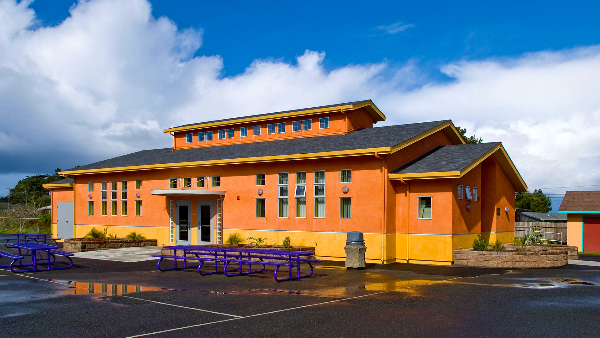 Exterior of completed cafeteria. showing asphalt and purple lunch tables, clouds and blue sky above.