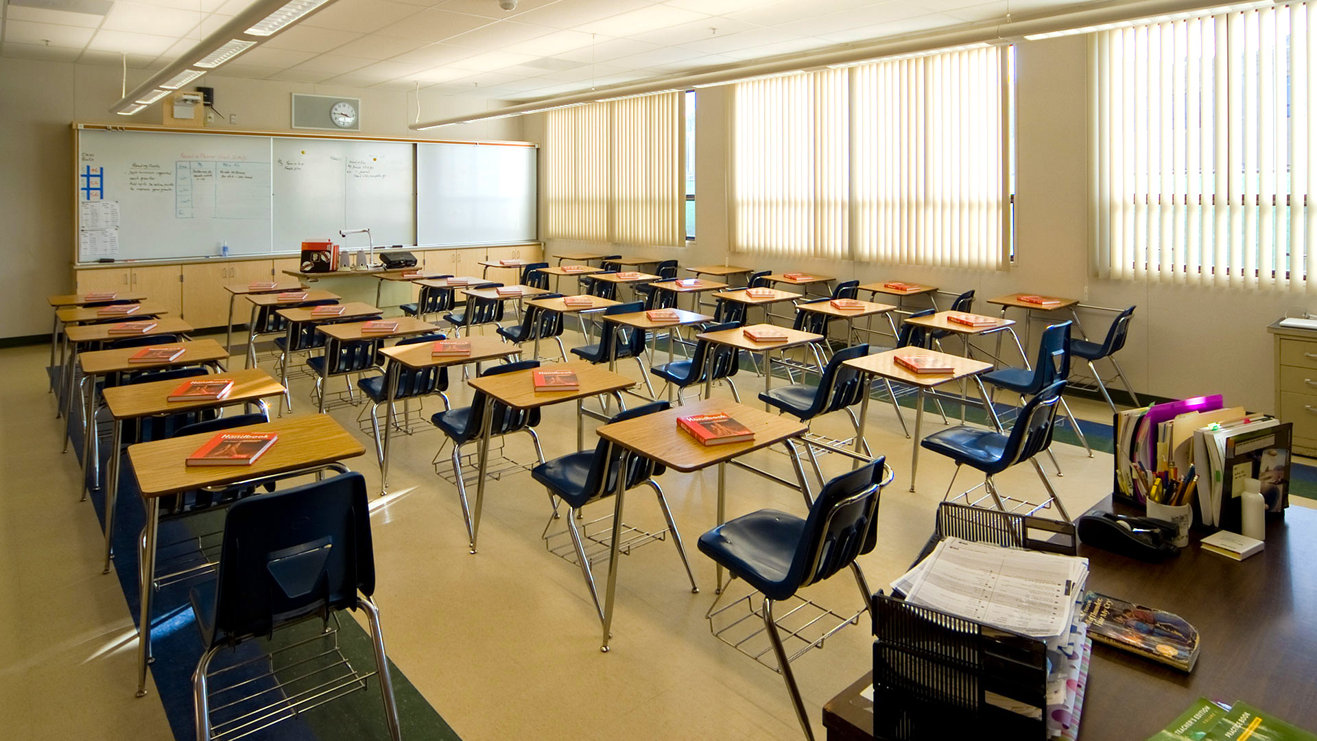 Classroom interior, with large, bright windows and blinds, and lots of desks focused towards teaching area.