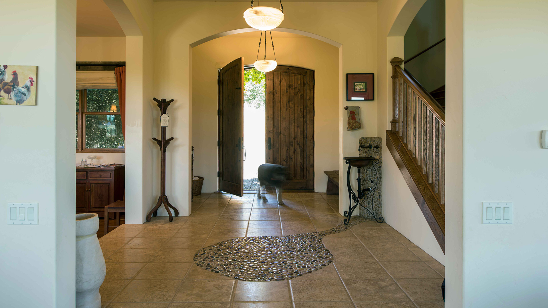 Interior of foyer, floor with inset stone circle leading to metal sink. Dog entering large wooden front door.