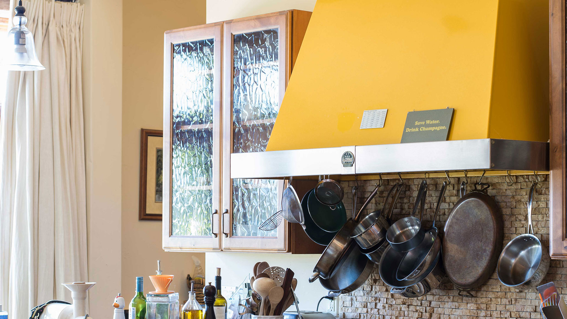 Closeup of yellow stove hood with pots hanging underneath. Wall cabinet with textures glass door behind.