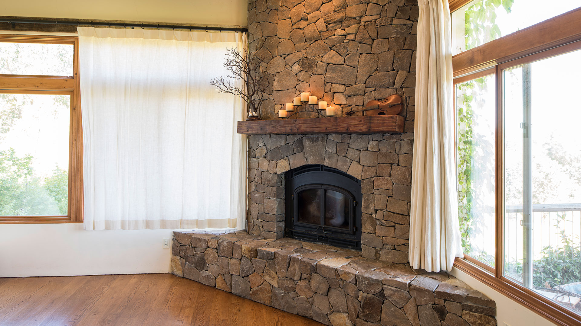 Interior of living room, stacked stone fireplace in corner framed with two large windows behind white drapes.