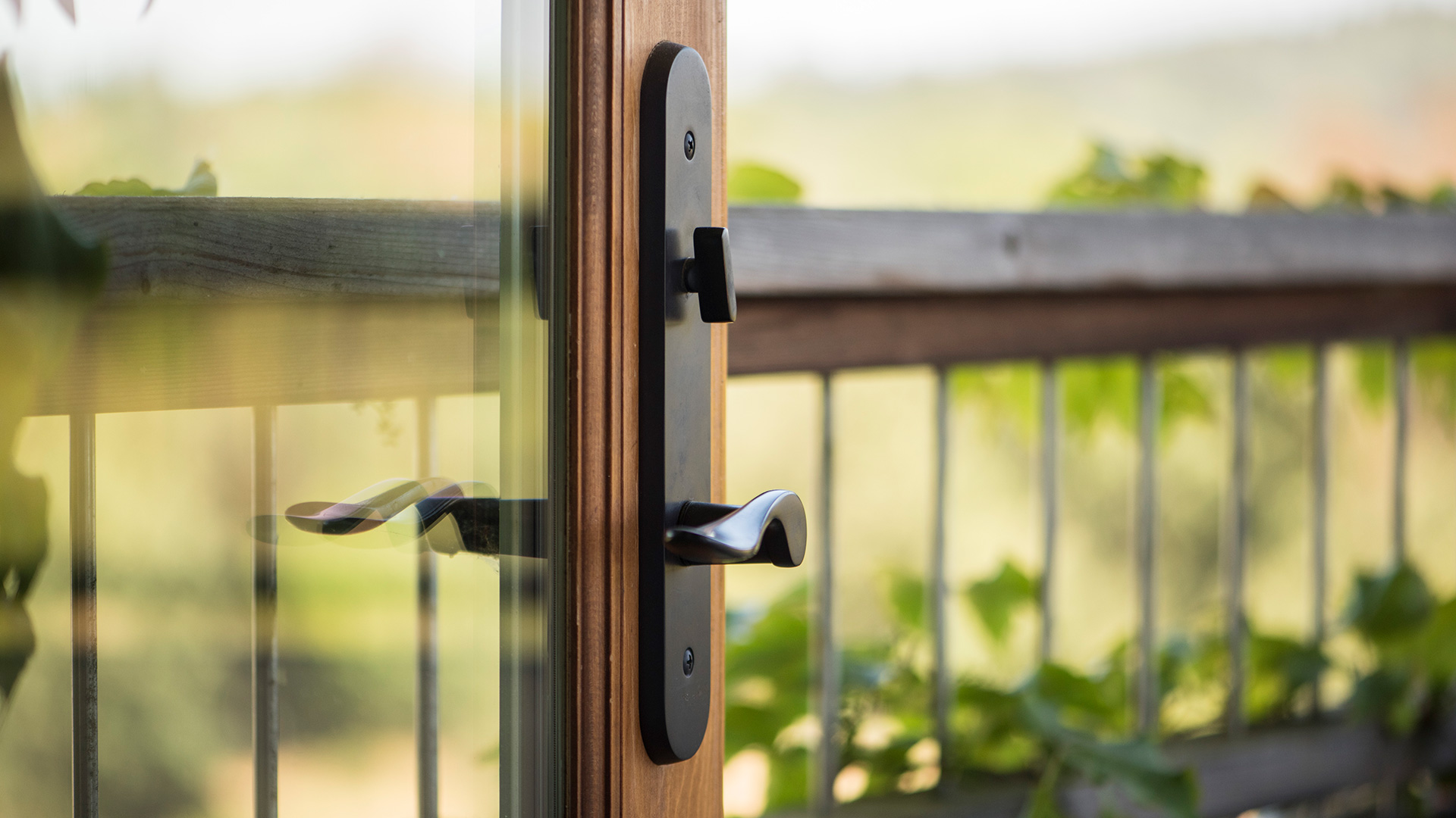 Closeup of handle on wooden frame of glass door, railing and plants reflected in door window.