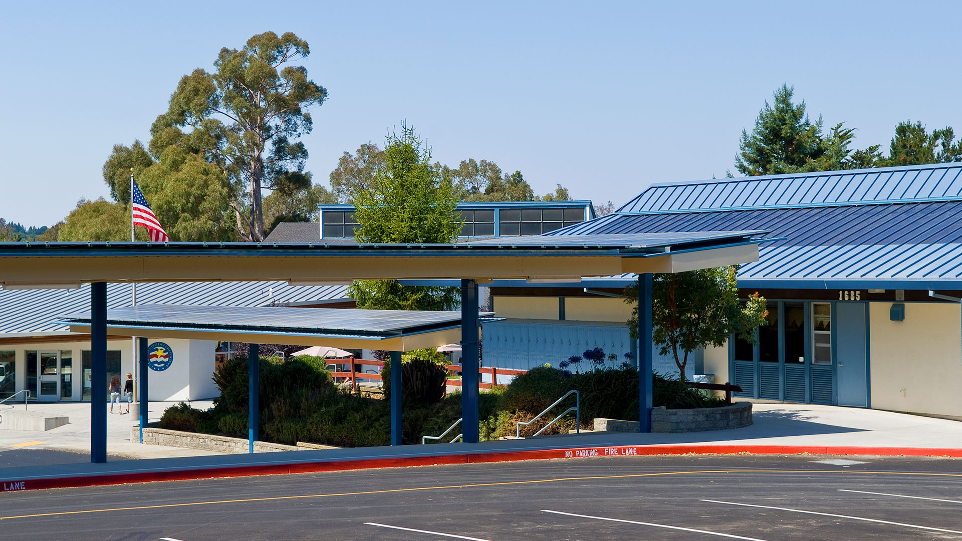 Covered walkways in parking area with solar panels extending along the roof. School campus behind.