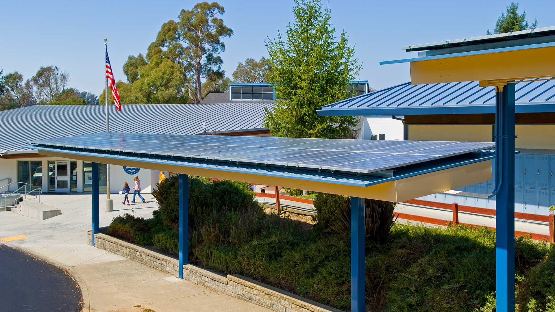 Covered walkways in parking area with solar panels extending along the roof. School campus behind.