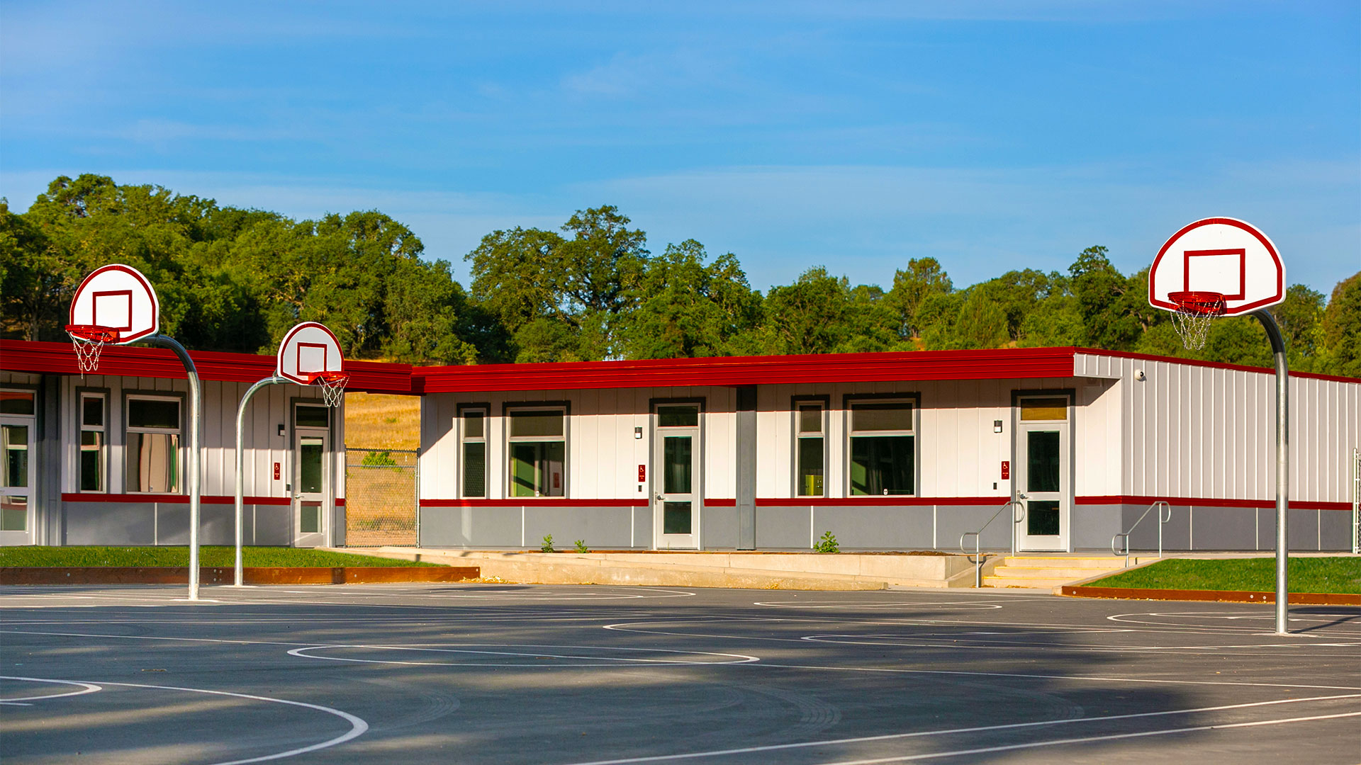 Portable classrooms with white panel walls, gray wainscot, and red trim. Blacktop with planters in front of building.