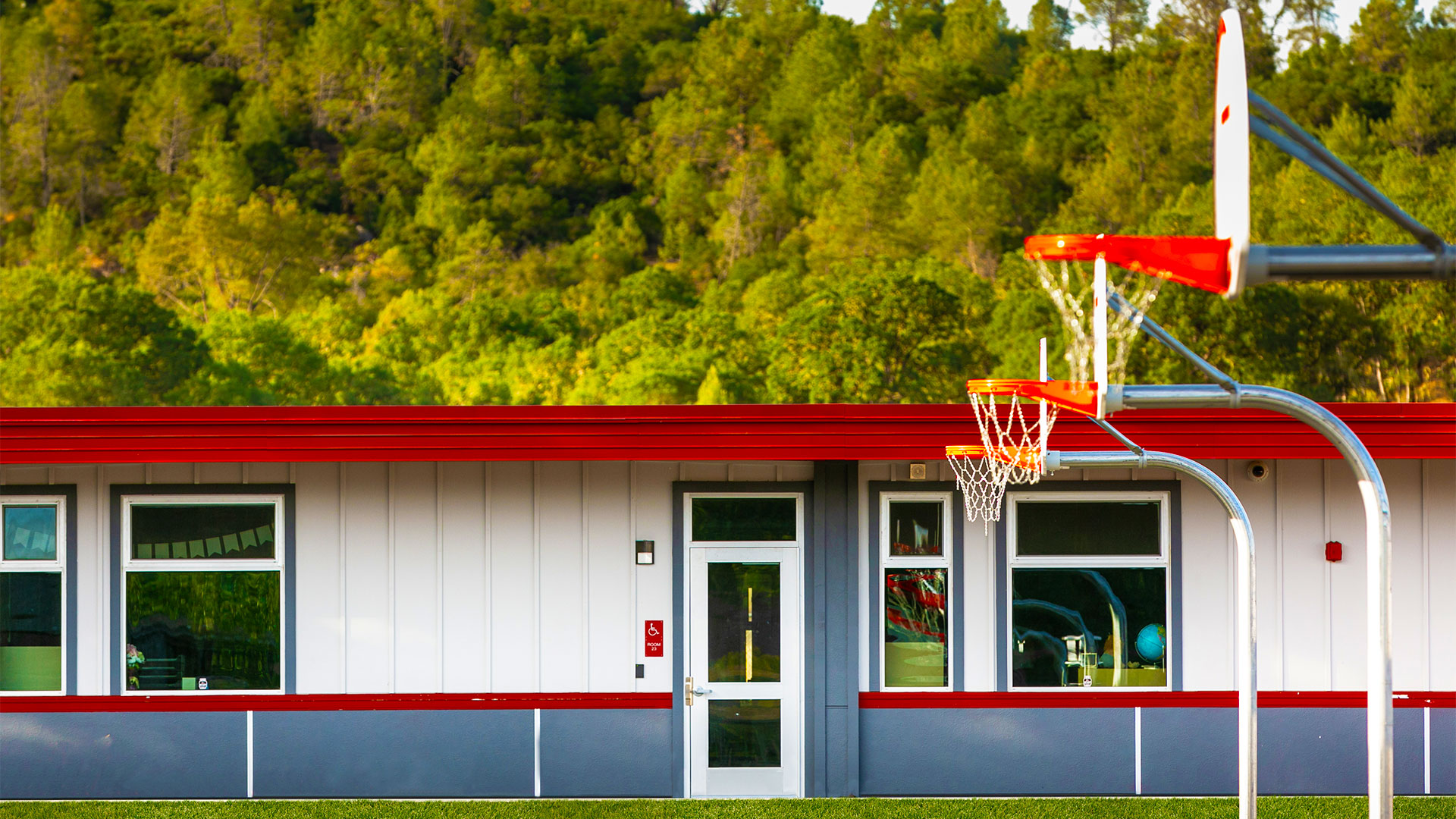 Portable classrooms with white panel walls, gray wainscot, and red trim. Basketball hoop in foreground and trees in back