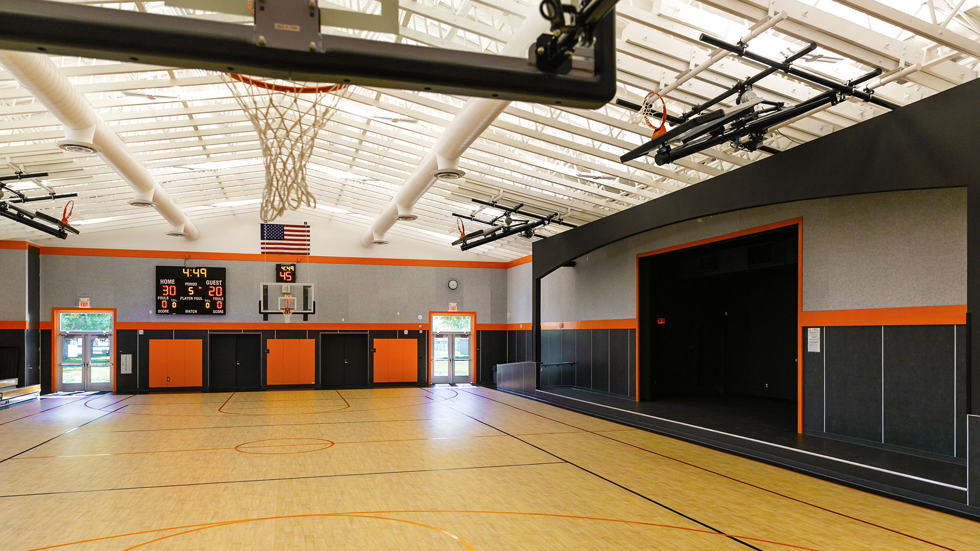 Interior of gym. Gray walls, black wainscot, and orange trim. Basketball court under white roof and trusses.