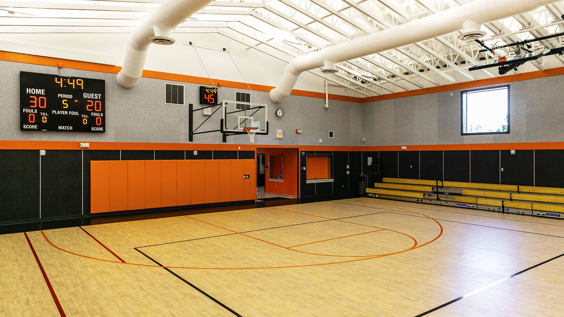 Interior of gym, looking at snack shack area with small bleachers adjacent. Shot clock above basketball hoop.