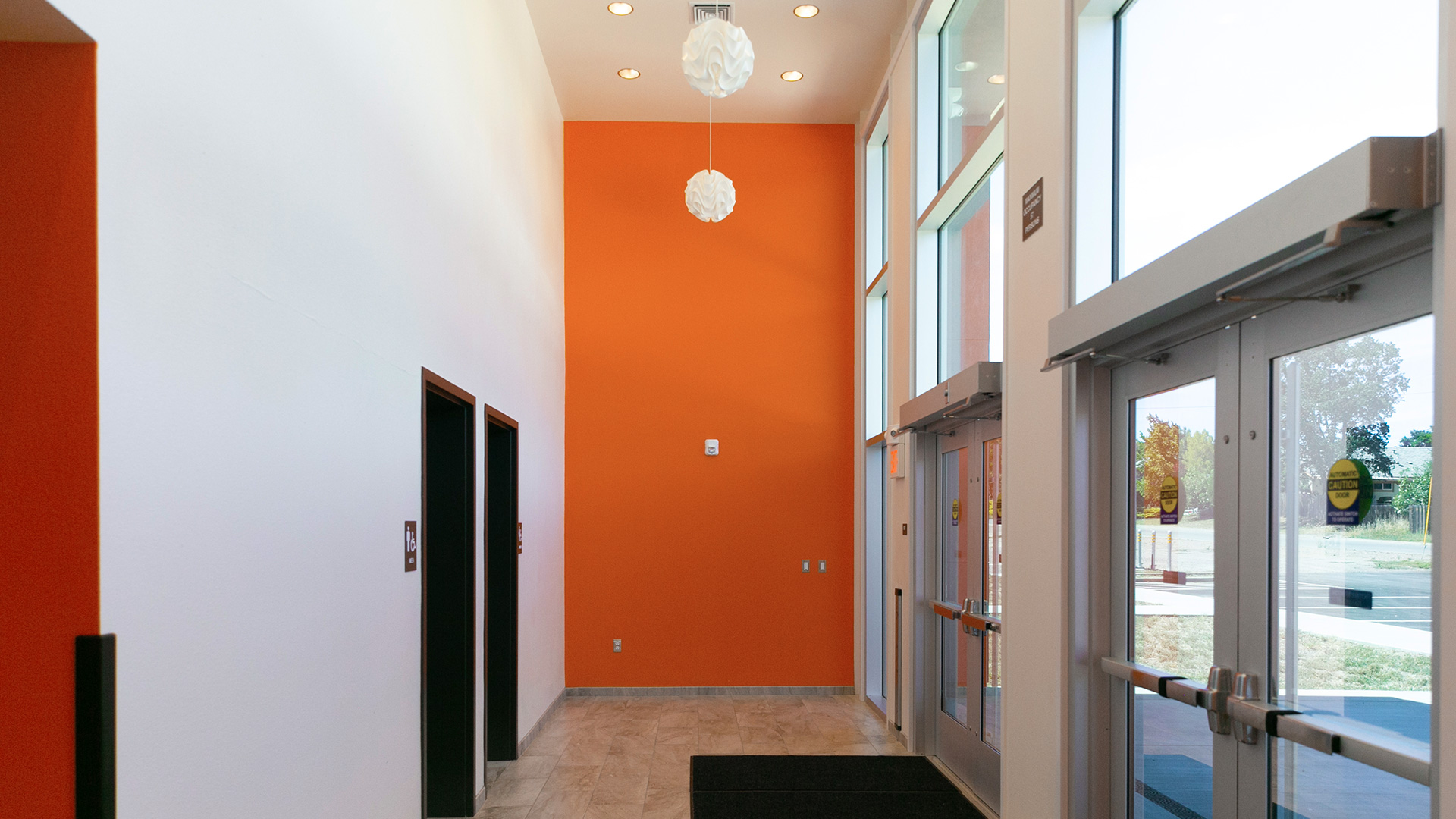 Interior of foyer with white wall and orange accent wall. Tall glass windows doors opposite bathroom entrances.