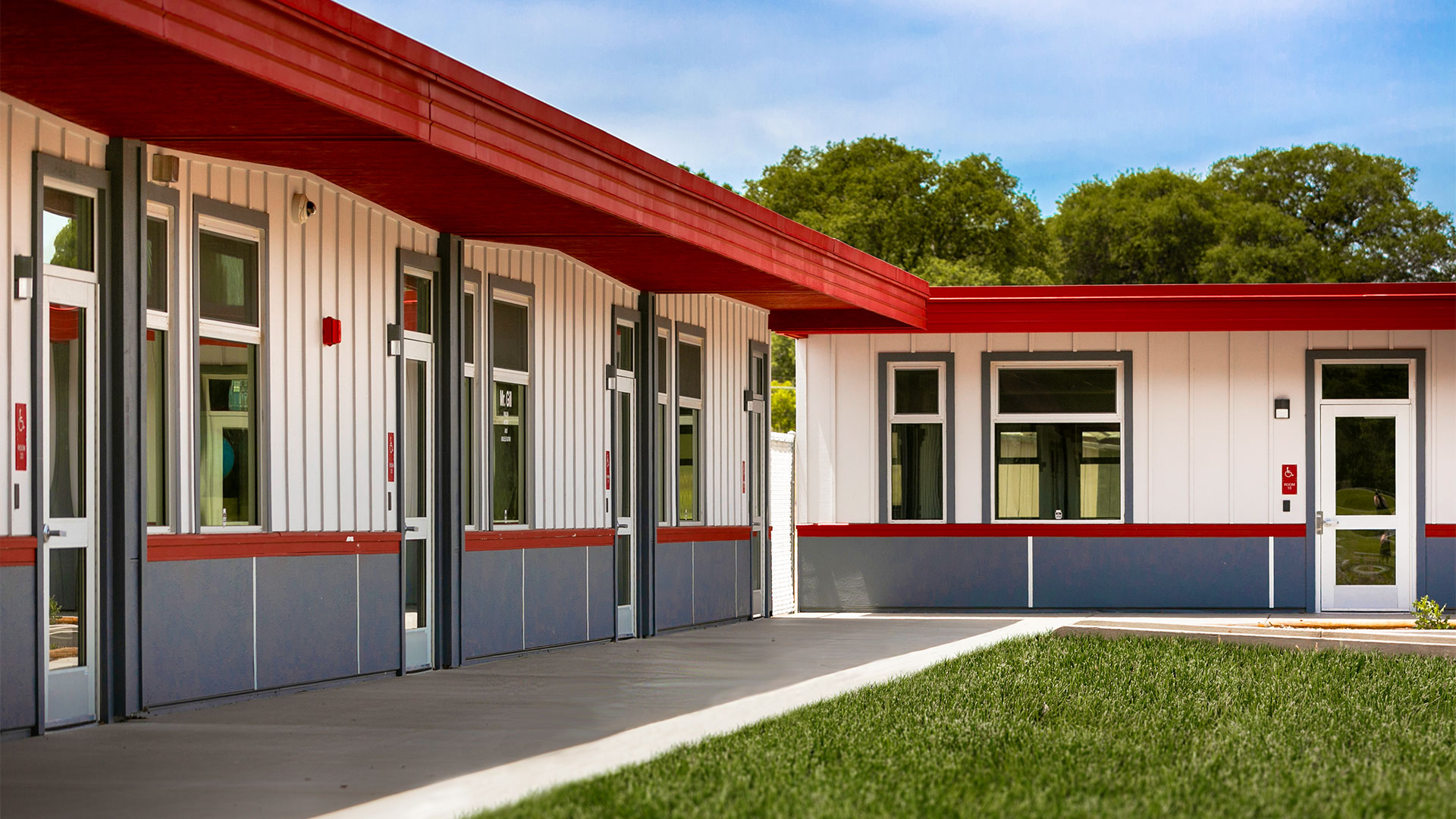 Portable classroom wing with white panel walls and gray wainscot, with concrete walkway and grass.
