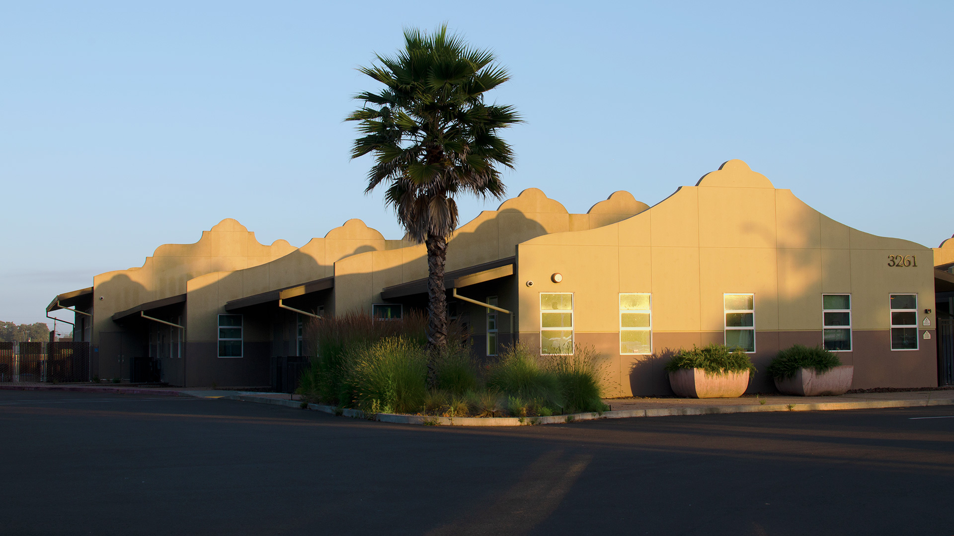 Entrance of school, with mission-style rooflines, beige walls, and gray wainscot. Bushes and palm tree landscaping.