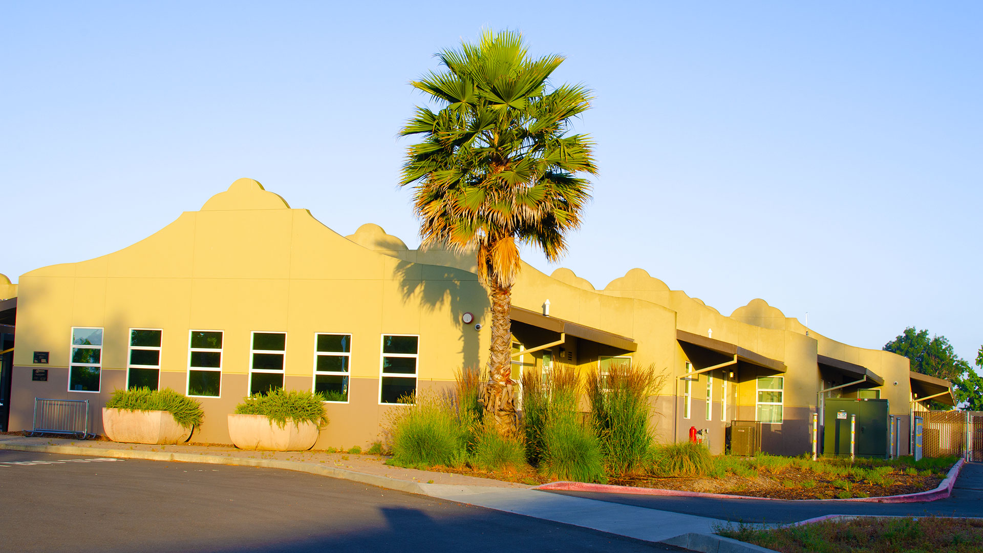 Entrance of school, with mission-style rooflines, beige walls, and gray wainscot. Bushes and palm tree landscaping.
