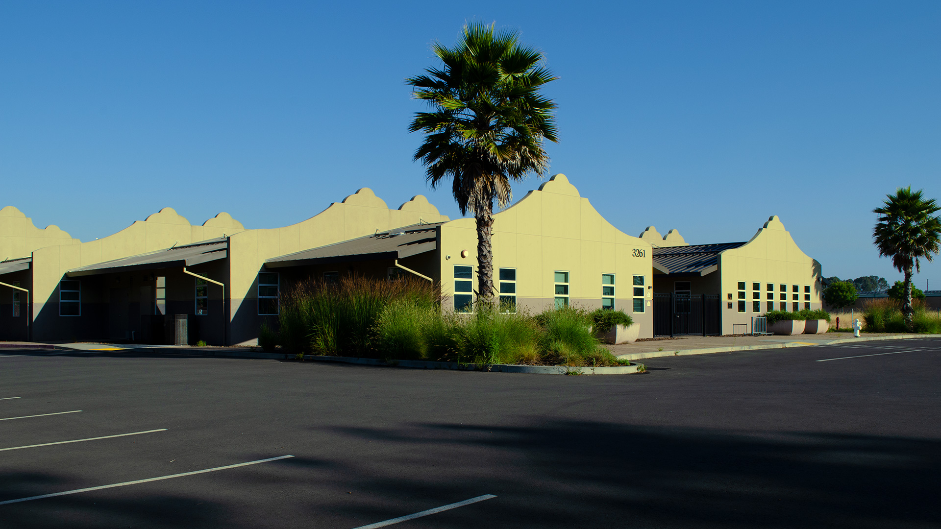 Entrance of school, with mission-style rooflines, beige walls, and gray wainscot. Bushes and palm tree landscaping.