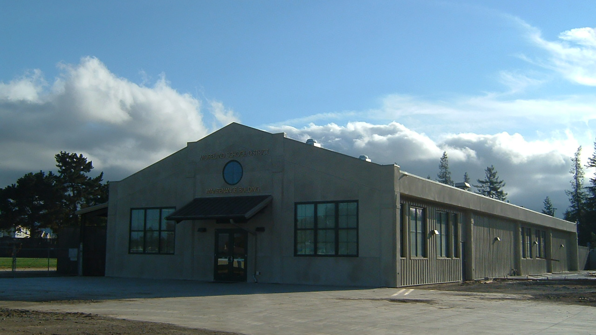 Exterior of concrete maintenance building with gable roof and circular window centered at wall peak.