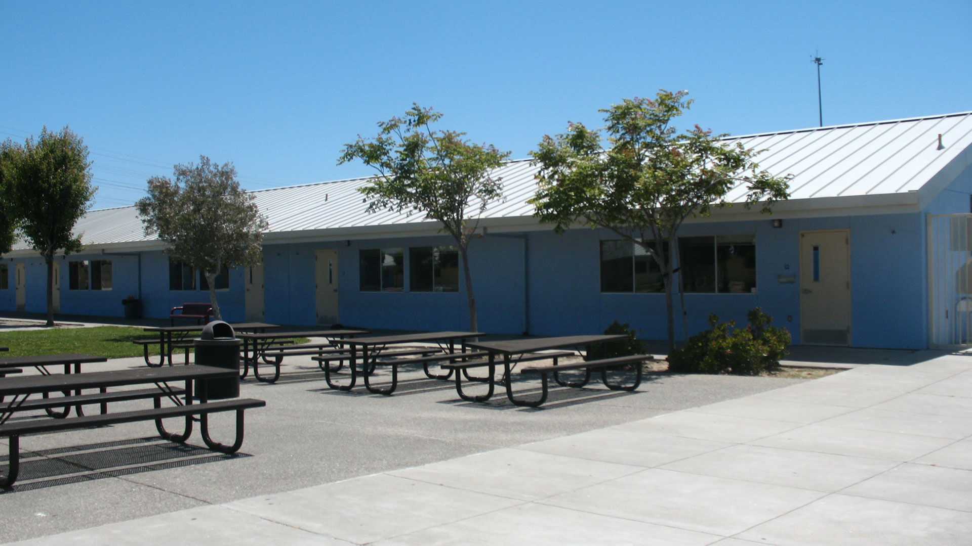 School courtyard with blue classroom buildings behind and lunch tables in front.