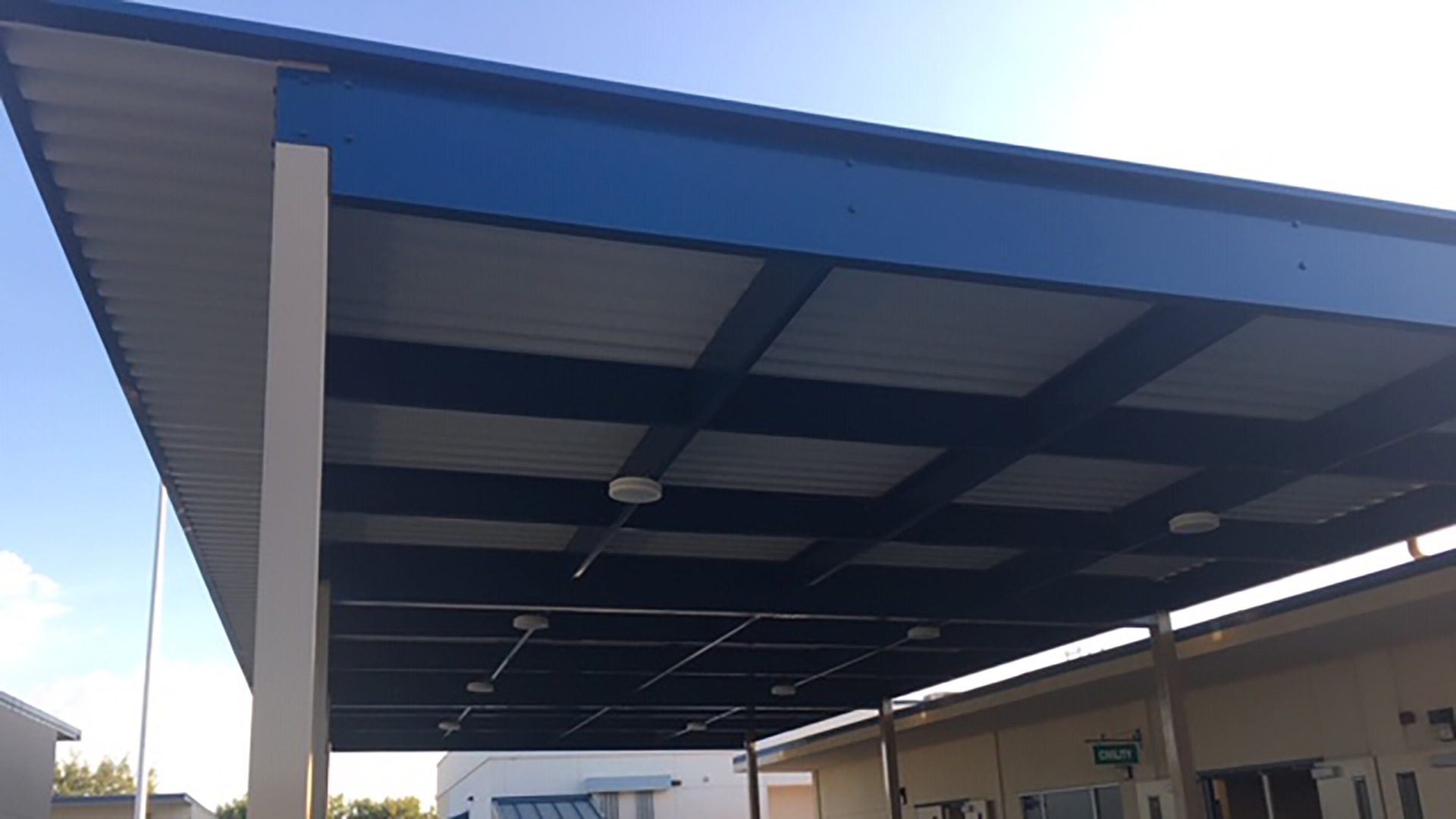 Large shade structure over concrete area between classrooms, with white posts and roof, and blue supports.