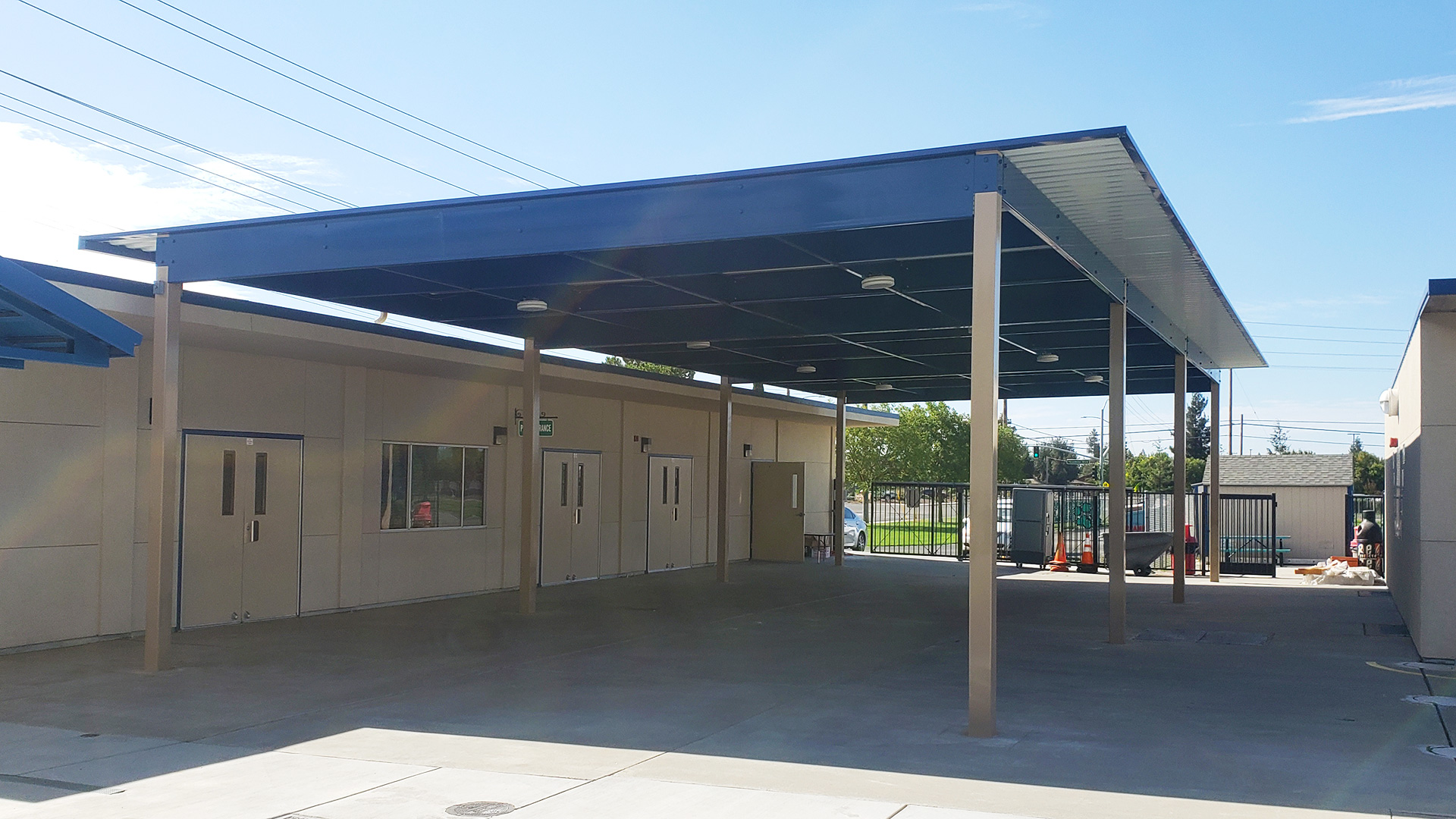 Large shade structure over concrete area between classrooms, with white posts and roof, and blue supports.