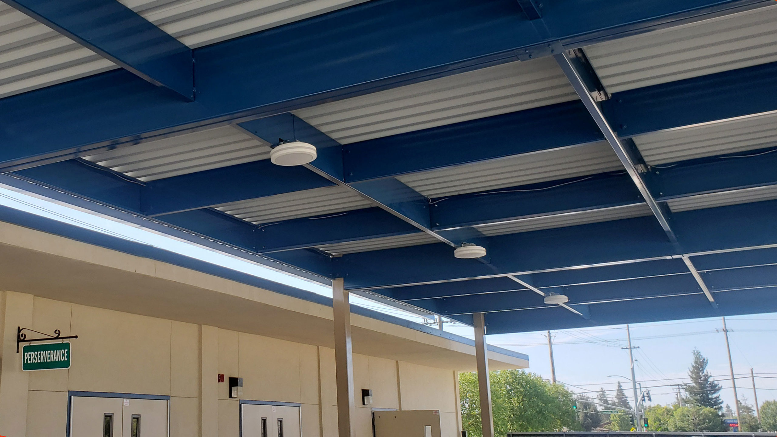 Large shade structure over concrete area between classrooms, with white posts and roof, and blue supports.