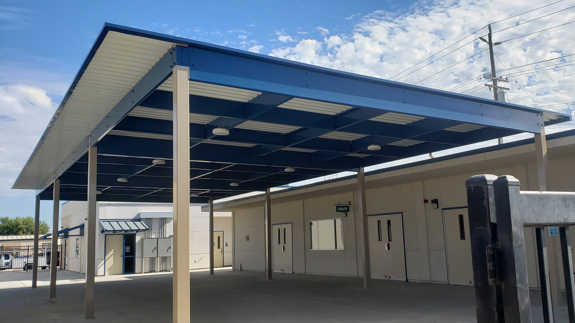 Large shade structure over concrete area between classrooms, with white posts and roof, and blue supports.